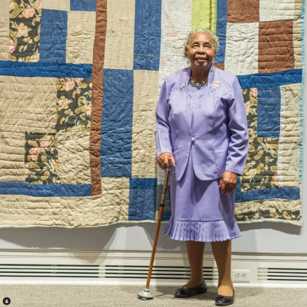 A woman in a purple skirt suit standing in front of a quilt that is mounted on a wall